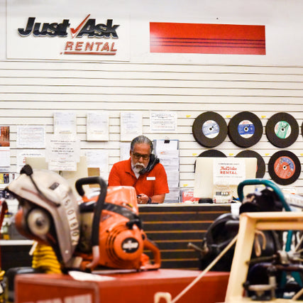 General RentalsEmployee in a red shirt assisting a customer at the rental counter of a hardware store, surrounded by various power tools and equipment with a 'Just Ask Rental' sign displayed on the wall.